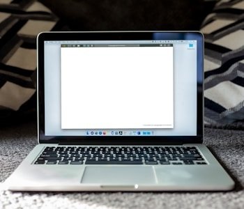 An open laptop on a carpeted surface displays the Gmail loading screen. The background is blurred, with patterned pillows or cushions adding a cozy setting.